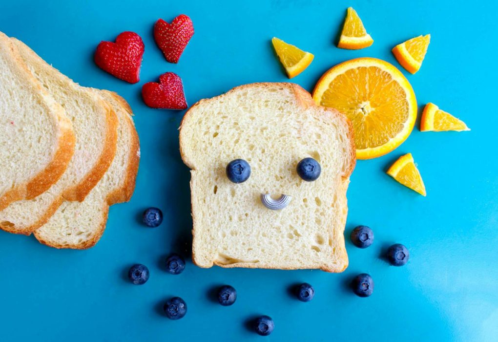 Fun and colorful toast face art with blueberries, strawberries, and orange slices on a blue background.