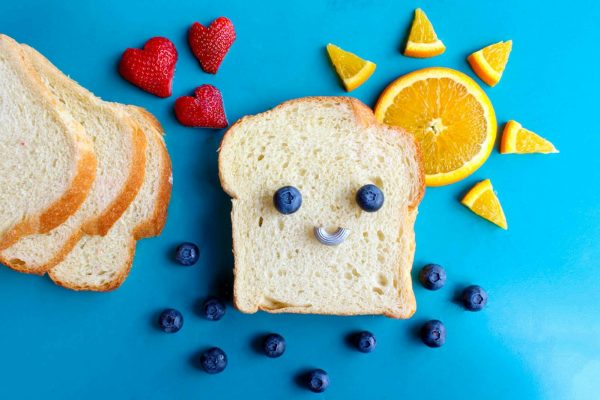 Fun and colorful toast face art with blueberries, strawberries, and orange slices on a blue background.