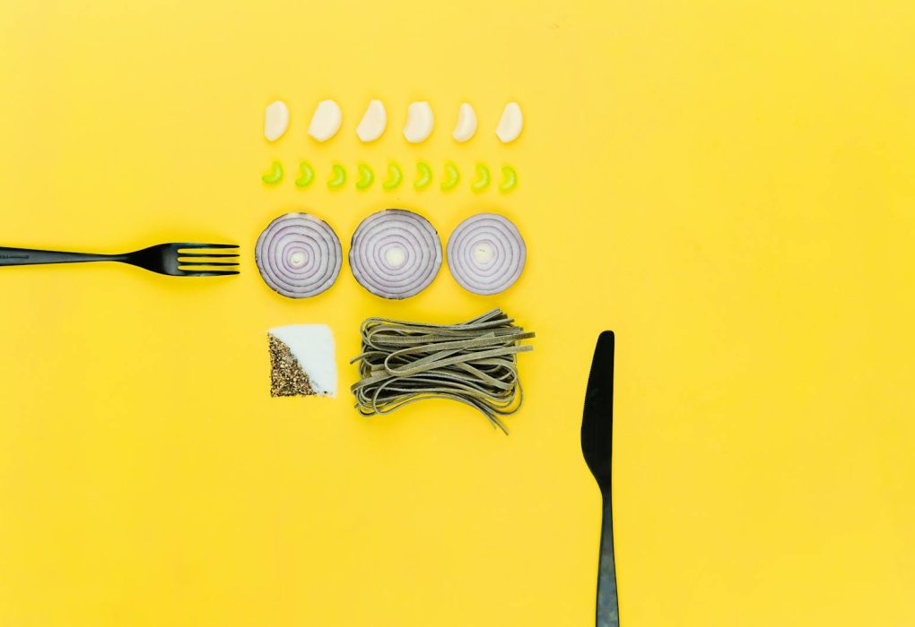 A colorful flatlay of pasta, onion, garlic, herbs, and cutlery on a yellow background.