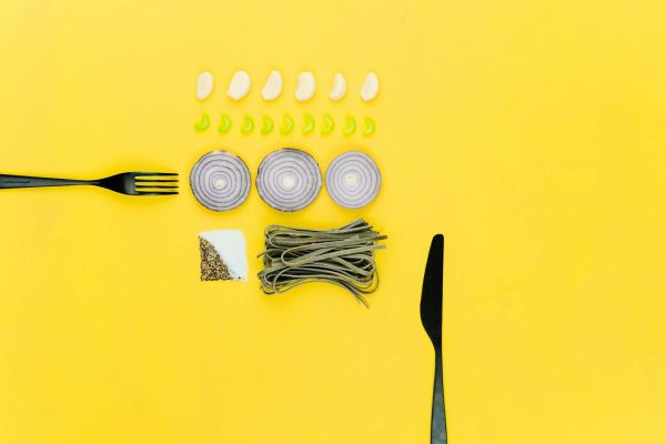 A colorful flatlay of pasta, onion, garlic, herbs, and cutlery on a yellow background.